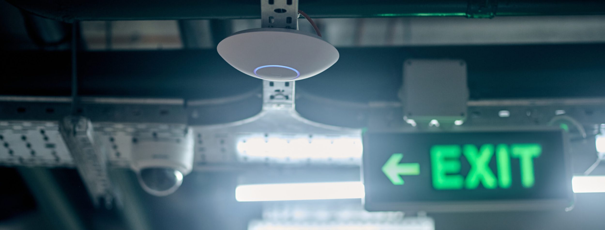 Optical smoke detector mounted on the ceiling beside the illuminated exit sign with a green arrow and the surveillance camera